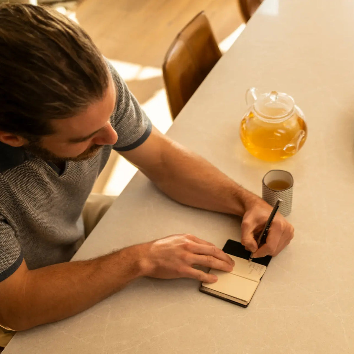 Man sitting at a table writing in a notebook with a teapot and cup in the background.