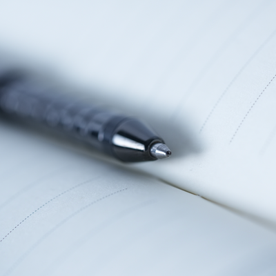A close-up, shallow depth-of-field shot of a black ballpoint pen resting on the pages of an open lined notebook.